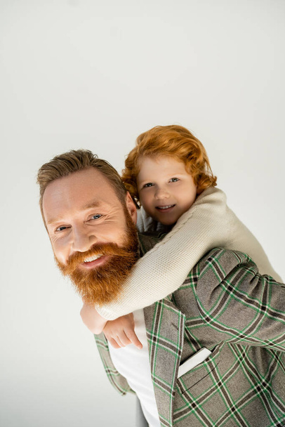 Smiling redhead boy in knitted jumper hugging bearded dad isolated on grey  - 写真・画像