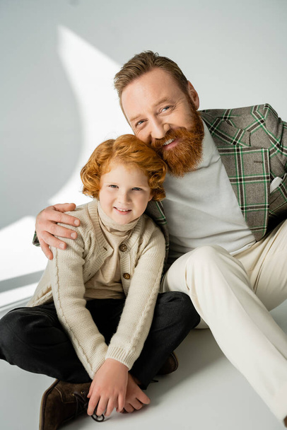 Wesoły brodaty mężczyzna przytulający rudego syna na szarym tle z jasnym  - Zdjęcie, obraz