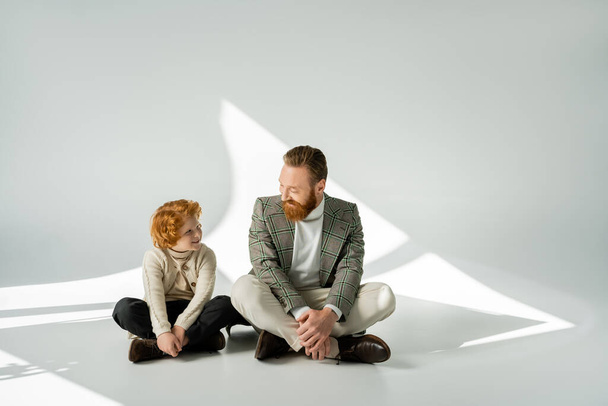 Cheerful redhead boy in knitted jumper looking at dad on grey background with light - Photo, Image
