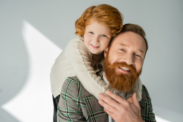 Redhead γιος σε ζεστό άλτης κοιτάζοντας κάμερα, ενώ αγκαλιάζει τον μπαμπά σε γκρι φόντο με φως  - Φωτογραφία, εικόνα