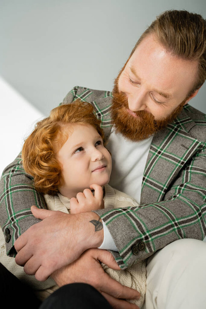 Happy man in checkered jacket hugging and looking at red haired boy on grey background - Fotoğraf, Görsel