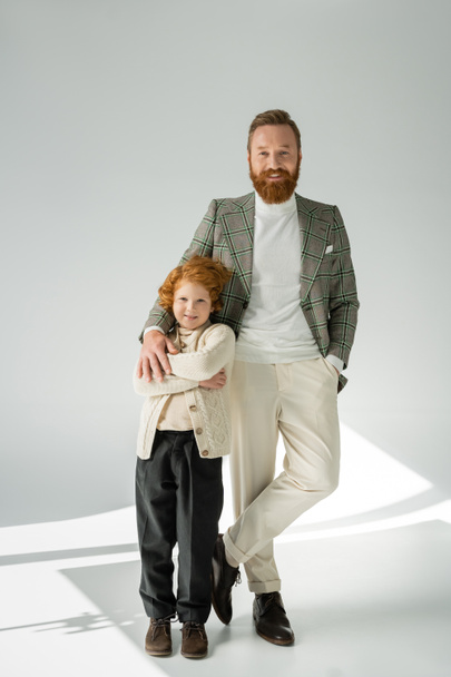 Full length of stylish father and redhead son posing and looking at camera on grey background with light  - 写真・画像
