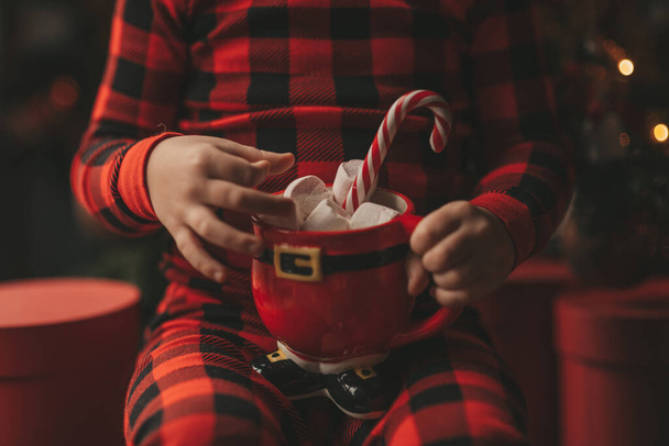 Happy little boy dreamer waiting at miracle Santa enjoy sweets color lollipop. Red checkered sleepwear cheerful kid celebrating new year hold Xmas cup with marshmallows near noel tree eve 25 december - Foto, afbeelding
