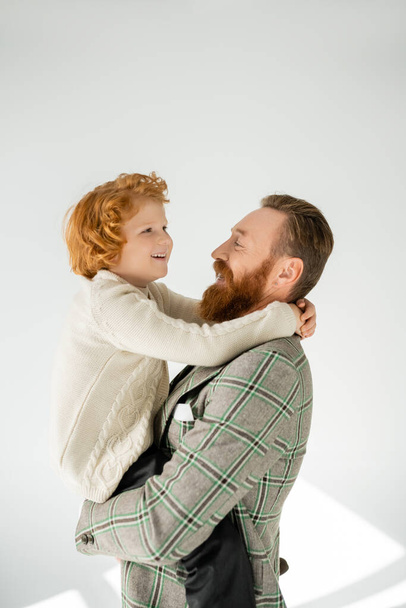 Trendy father in jacket hugging and lifting cheerful redhead son on grey background - Photo, Image
