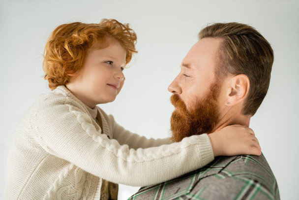 Smiling red haired child hugging bearded father in jacket isolated on grey  - Fotografie, Obrázek