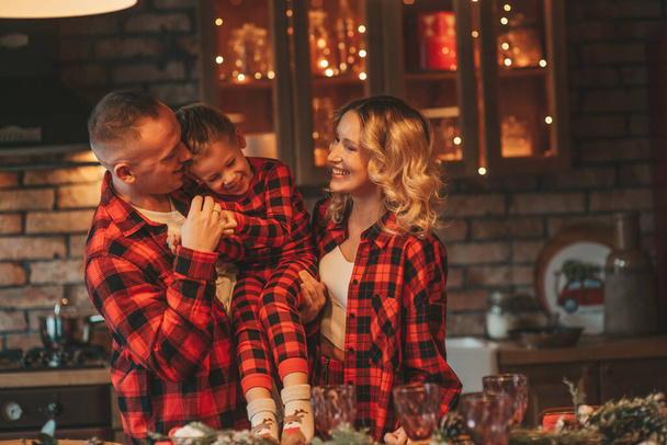 Smile active parents with small son in red checkered sleepwears waiting Santa indoor. Stylish family love laughing hugs kisses at home. Celebrate New year garlands and lights eve 25 December Noel tree - 写真・画像