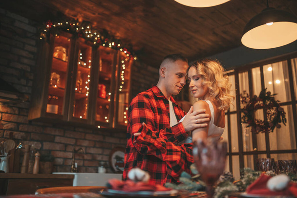 Portrait of young lovely couple hugging indoor eve 25 December. Lovers laughing hugs kisses waiting miracle at home. Celebrating new year garlands lights noel in red plaid pajamas passion tenderness - Photo, image