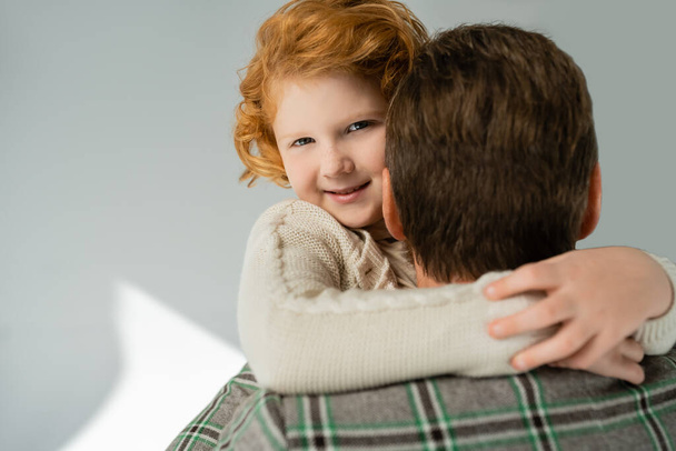 Cheerful red haired boy in knitted jumper hugging blurred dad on grey background - Foto, Imagen