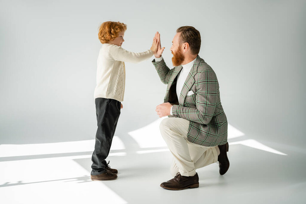 Πλευρική άποψη του κοκκινομάλλα αγόρι δίνοντας κόλλα πέντε σε κομψό πατέρα σε γκρι φόντο με φως  - Φωτογραφία, εικόνα