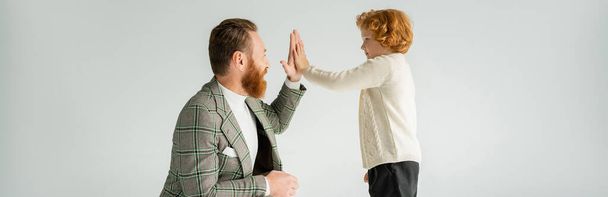 Side view of smiling bearded father giving high five to red haired son isolated on grey, banner  - Photo, Image
