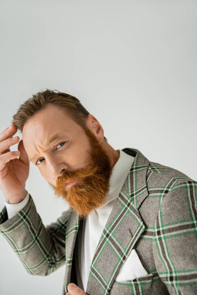 Trendy bearded man in jacket touching hair isolated on grey  - Fotografie, Obrázek