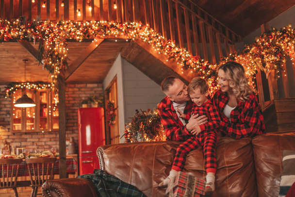 Smile active parents with small son in red checkered sleepwears waiting Santa indoor. Stylish family love laughing hugs kisses at home. Celebrate New year garlands and lights eve 25 December Noel tree - Photo, Image