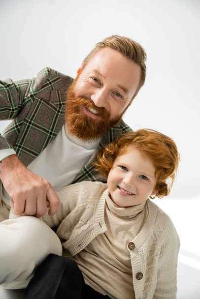 Portrait of trendy father and redhead boy smiling at camera on grey background with light  - Photo, Image