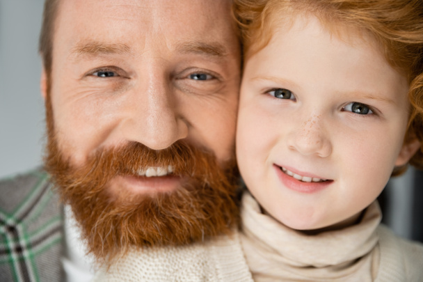 Portrait of redhead boy smiling at camera near bearded dad on grey background - Photo, Image