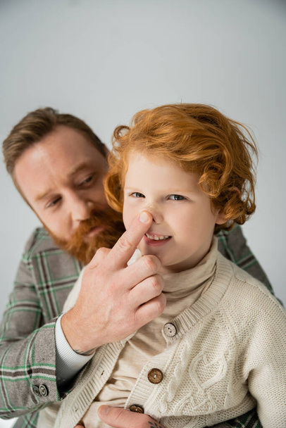 Blurred dad in jacket touching nose of red haired son isolated on grey  - Foto, immagini