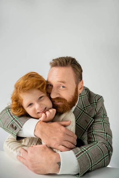 Smiling man in plaid jacket hugging red haired son and looking at camera on grey background - Foto, afbeelding