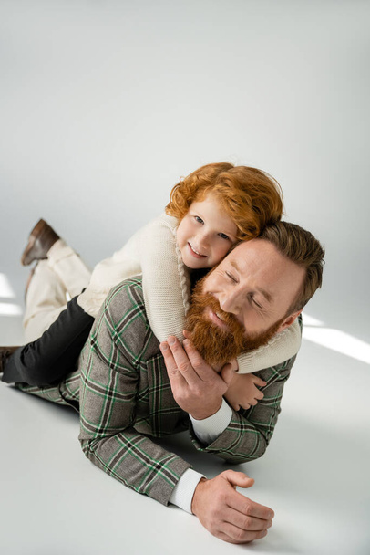 Redhead child in jumper hugging cheerful dad while lying on grey background - Photo, Image