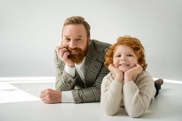 cheerful redhead father and son in stylish clothes lying and looking at camera on grey background - Photo, Image