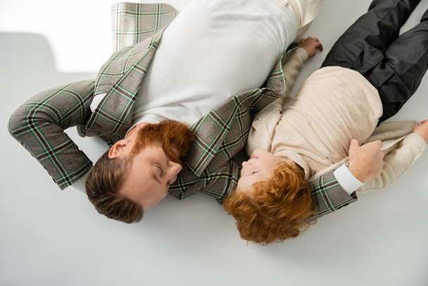 top view of redhead father and son in trendy outfit looking at each other while lying on grey background - Photo, Image