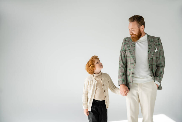 redhead father and son in trendy outfit holding hands and looking at each other on grey background - Zdjęcie, obraz