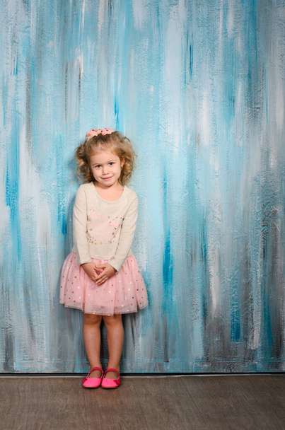 a beautiful girl in a light jacket and a pink tutu skirt and shoes stands against a blue wall in a watercolor style - Photo, Image