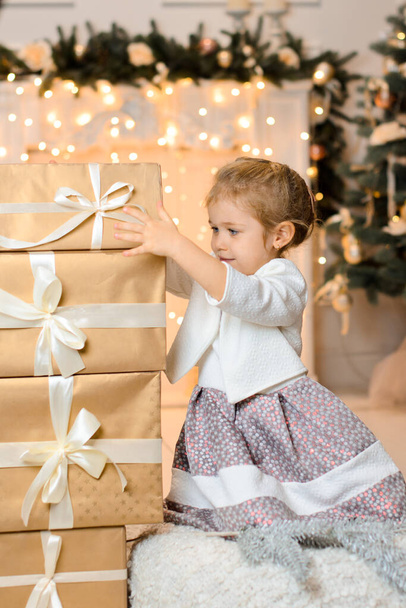 piękna szczęśliwa dziewczyna siedzi na podłodze, na tle świątecznego kominka z lampkami girlandowymi, i umieszcza pudełka z prezentami na stosie - Zdjęcie, obraz