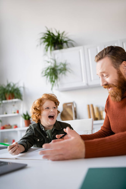 bearded man talking to laughing redhead son doing homework in kitchen - Foto, afbeelding