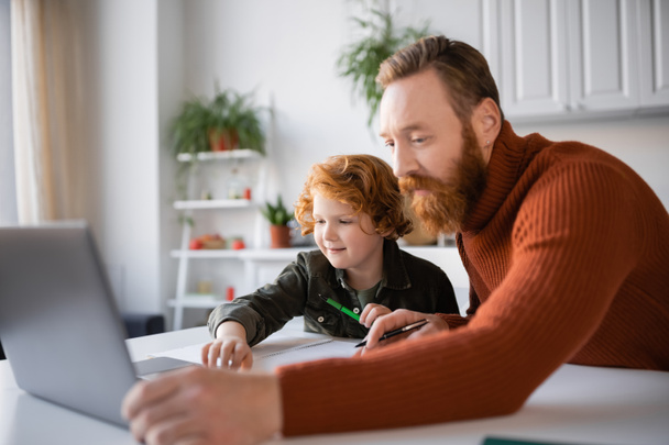 redhead father and son looking at blurred laptop while doing homework together - Photo, Image