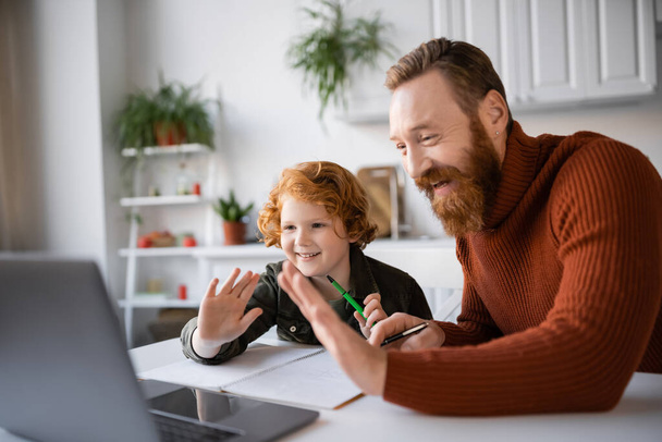 happy redhead father and son waving hands near laptop while doing homework together - Foto, immagini