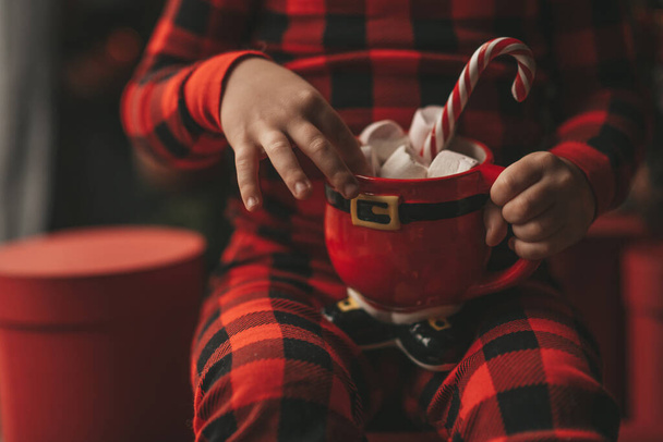 Šťastný chlapec snílek čeká na zázrak Santa vychutnat sladkosti barva lízátko. Červené kostkované spací oděv veselý dítě slaví Nový rok držet Vánoční pohár s marshmallows poblíž Noel tree Eve 25 Prosinec - Fotografie, Obrázek