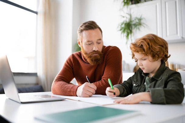 redhead child writing in notebook near bearded dad and laptop  - Photo, Image