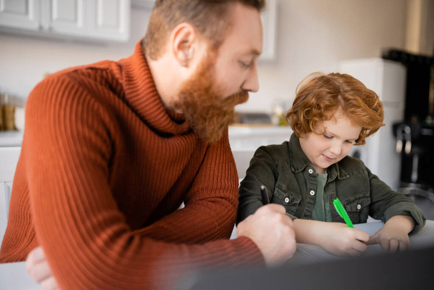 bearded man looking at redhead son writing in notebook while doing homework on blurred foreground - Фото, зображення