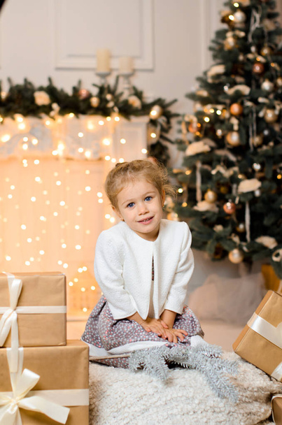 een mooi meisje in een witte trui zit in de buurt van geschenkdozen met een lint, tegen de achtergrond van een kerstboom, en een gloeiende bloemenslinger op de haard, bokeh lichten - Foto, afbeelding