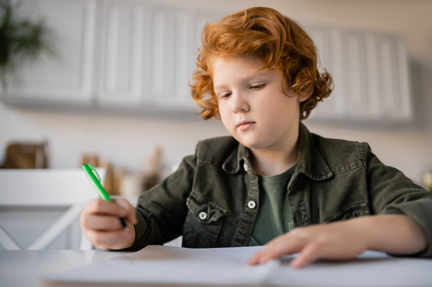 pensive redhead boy holding pen while doing homework near blurred notebook - Photo, Image