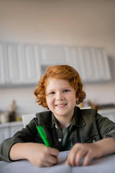 happy redhead kid with pen looking at camera while doing homework on blurred foreground - Fotó, kép