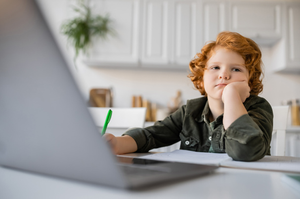 ennuyé rousse garçon assis avec la main près du visage tout en faisant des devoirs à l'ordinateur portable flou - Photo, image