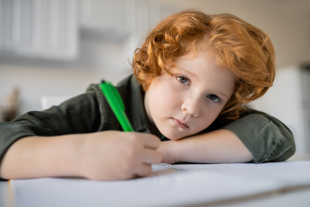 znudzony rudy chłopiec z długopisem patrząc na kamerę w pobliżu rozmazanego notatnika - Zdjęcie, obraz