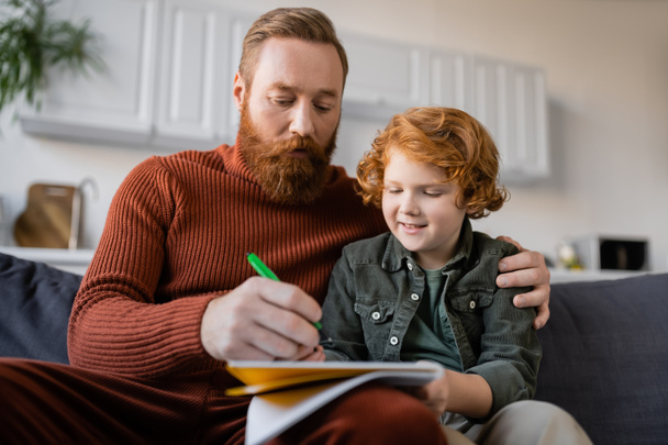 bearded man writing in notebook while sitting on sofa near smiling redhead son - Foto, afbeelding