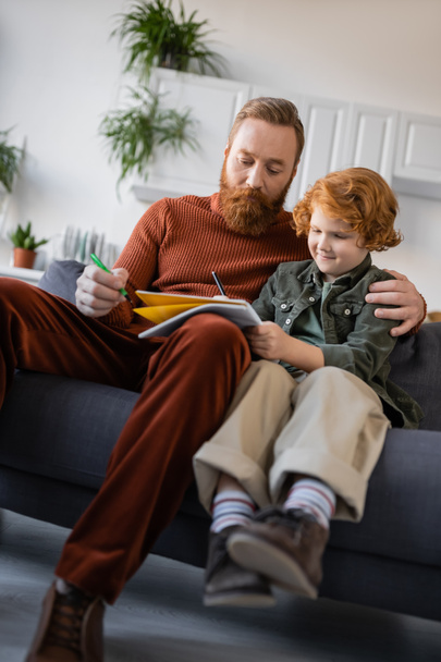redhead boy writing in copybook near bearded dad on couch at home - 写真・画像