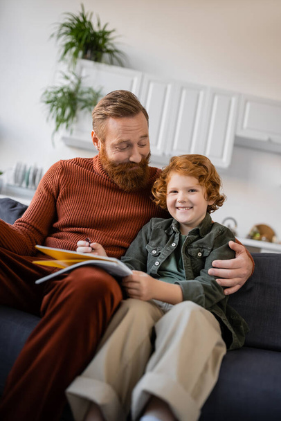 happy bearded man hugging cheerful redhead son sitting on couch with notebook - Fotografie, Obrázek