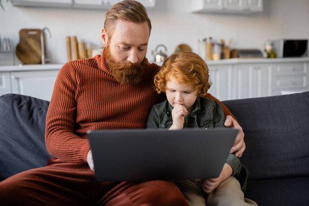 figyelmes vörös hajú gyermek nézi laptop, miközben ül a kanapén mellett szakállas apa - Fotó, kép