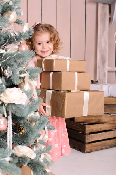 a beautiful smiling girl peeks out from behind a decorated Christmas tree, holds a stack of three boxes of gifts in her hands - Zdjęcie, obraz