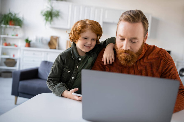 redhead kid smiling near bearded dad working on blurred laptop at home - Photo, Image