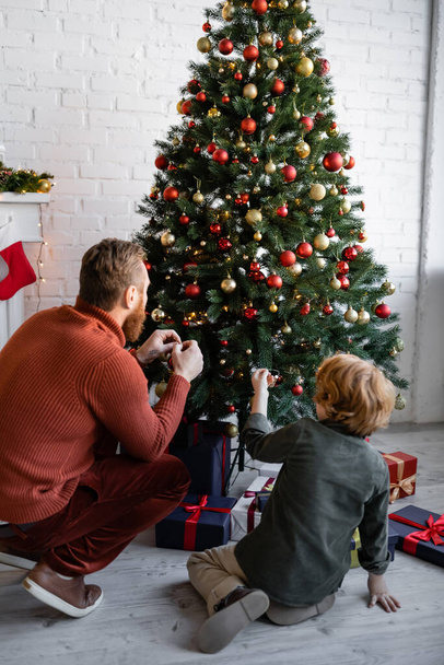 ギフトボックスの近くにクリスマスツリーを飾る赤毛の父と息子のバックビュー - 写真・画像