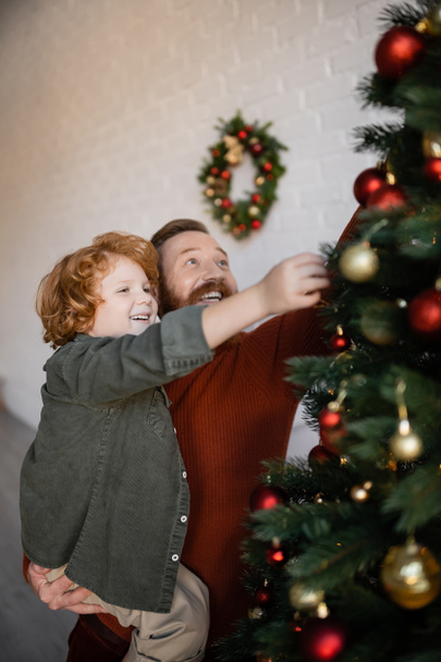 happy man with redhead son decorating Christmas tree on blurred foreground - Photo, Image