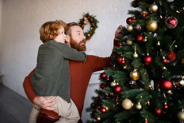 レッドヘッドの息子を抱えた髭の男がクリスマスツリーを家で飾り - 写真・画像