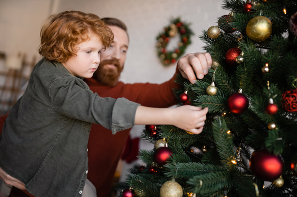 blurred man holding redhead son while decorating spruce with Christmas baubles at home - Photo, Image