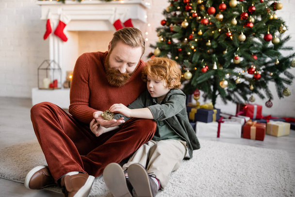 vörös hajú apa és a gyermek tartja karácsonyi csecsebecse a padlón a nappaliban ünnepi dekoráció - Fotó, kép