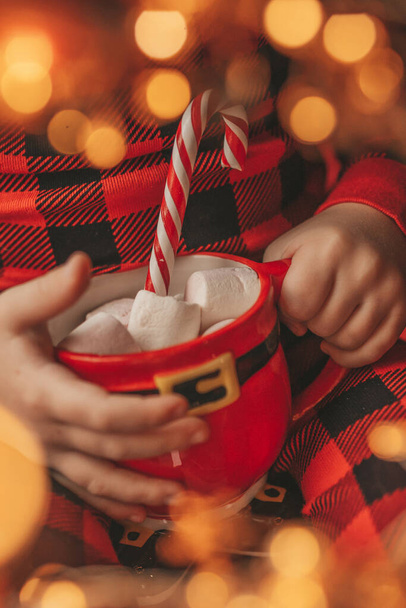 Счастливый маленький мальчик мечтатель ждет в чудо Санта наслаждаться сладостями цвета леденец. Красная клетчатая одежда для сна веселый ребенок празднует Новый год провести Рождество Кубок с зефиром рядом с Ноэль дерево канун 25 декабря - Фото, изображение