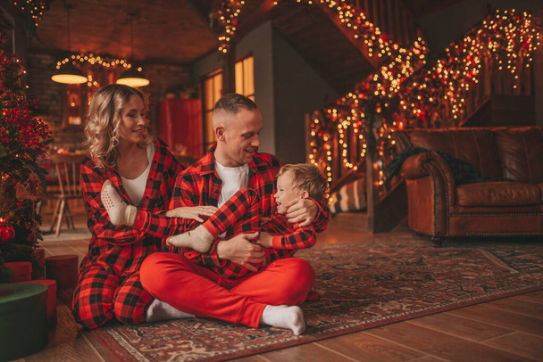 Uśmiechnij się aktywnych rodziców z małym synem w czerwonym kratkę śpiochy czeka Santa kryty. Stylowa rodzina uwielbia się przytulać do domu. Świętuj Nowy Rok girlandy i światła w wigilię 25 grudnia Noel tree - Zdjęcie, obraz